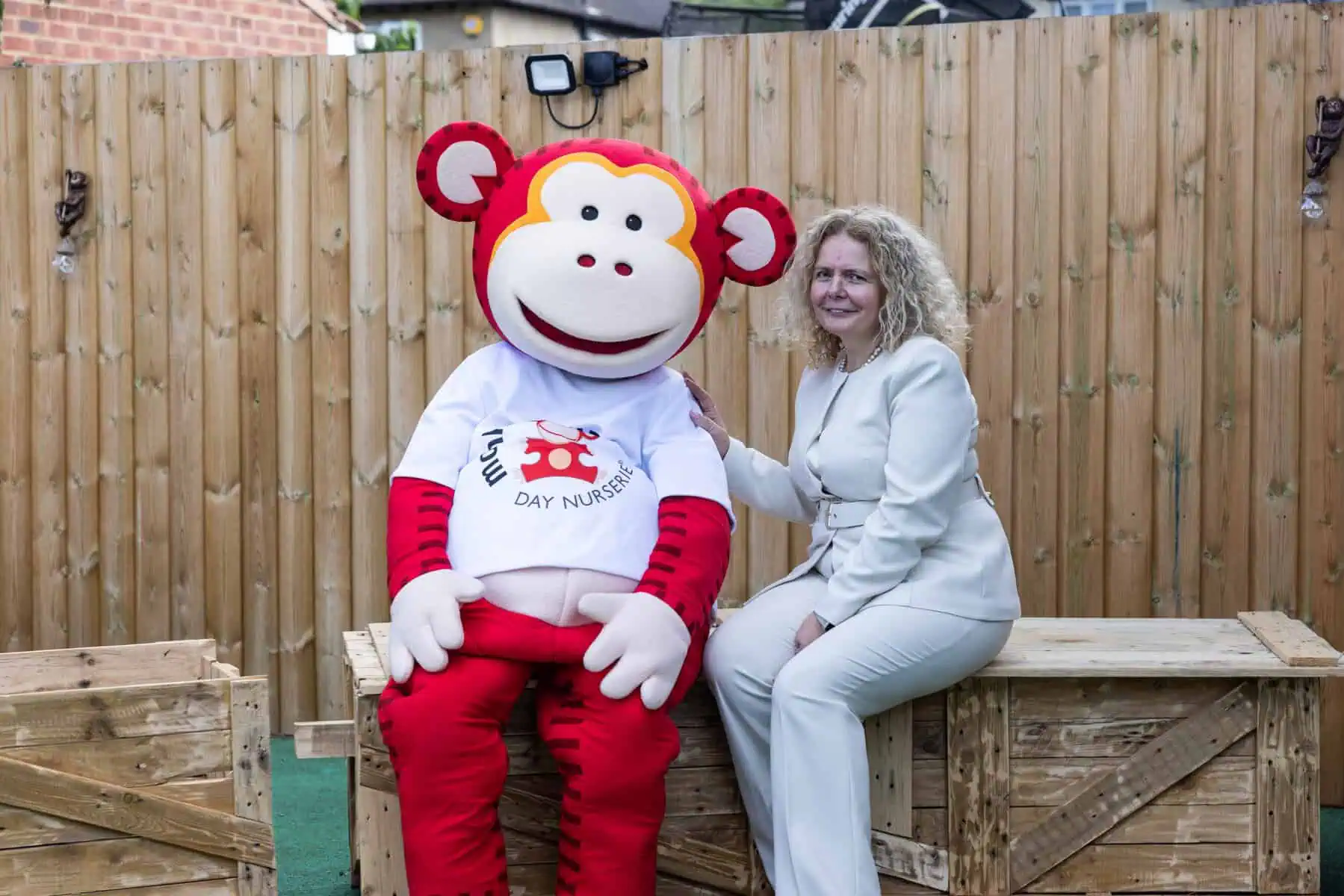 Monkey Puzzle Kingston Day Nursery Team Person in white suit with curly hair sitting next to a monkey mascot.